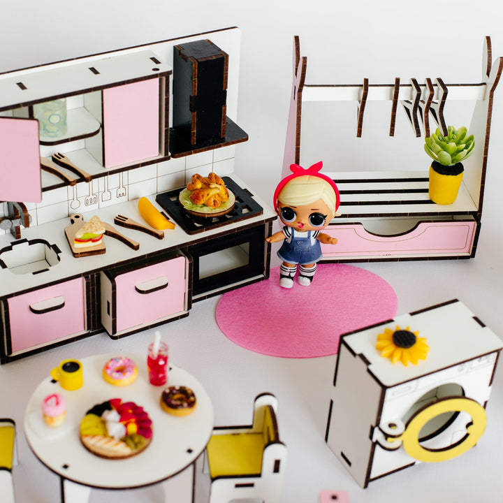 A miniature dollhouse kitchen and room set with pink and white furniture, a small doll standing on a pink rug, a dining table with toy food, a clothing rack, and a washing machine, all arranged on a white surface against a white background.