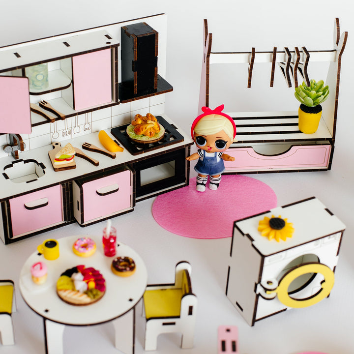A miniature dollhouse kitchen set with a small doll figure, including a pink and white kitchen cabinet, dining table with chairs, washing machine, clothes rack, and small accessories, arranged on a white surface against a white background.