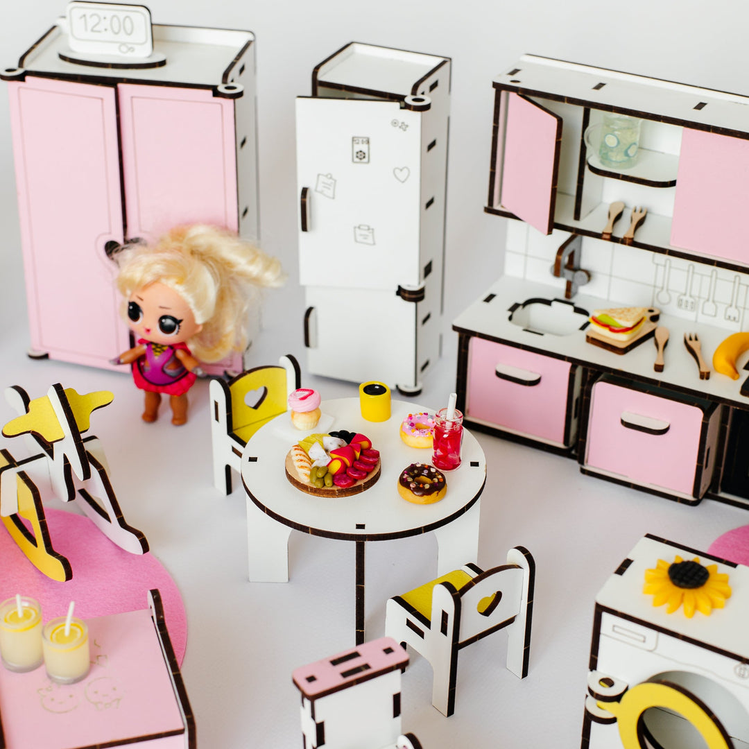 A miniature dollhouse-style play kitchen and dining set made from layered cardboard, featuring pink and white cabinets, a small round table with chairs, a refrigerator, washing machine, and a small doll standing beside the room.