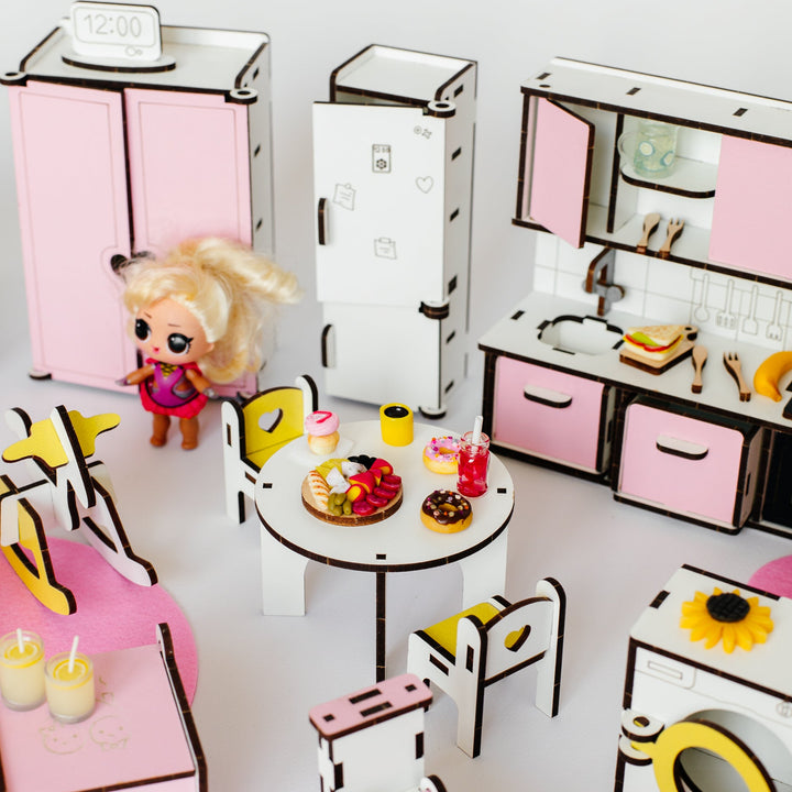 A miniature dollhouse-style play kitchen and dining set made from layered cardboard, featuring pink and white cabinets, a small round table with chairs, a refrigerator, washing machine, and a small doll standing beside the room.