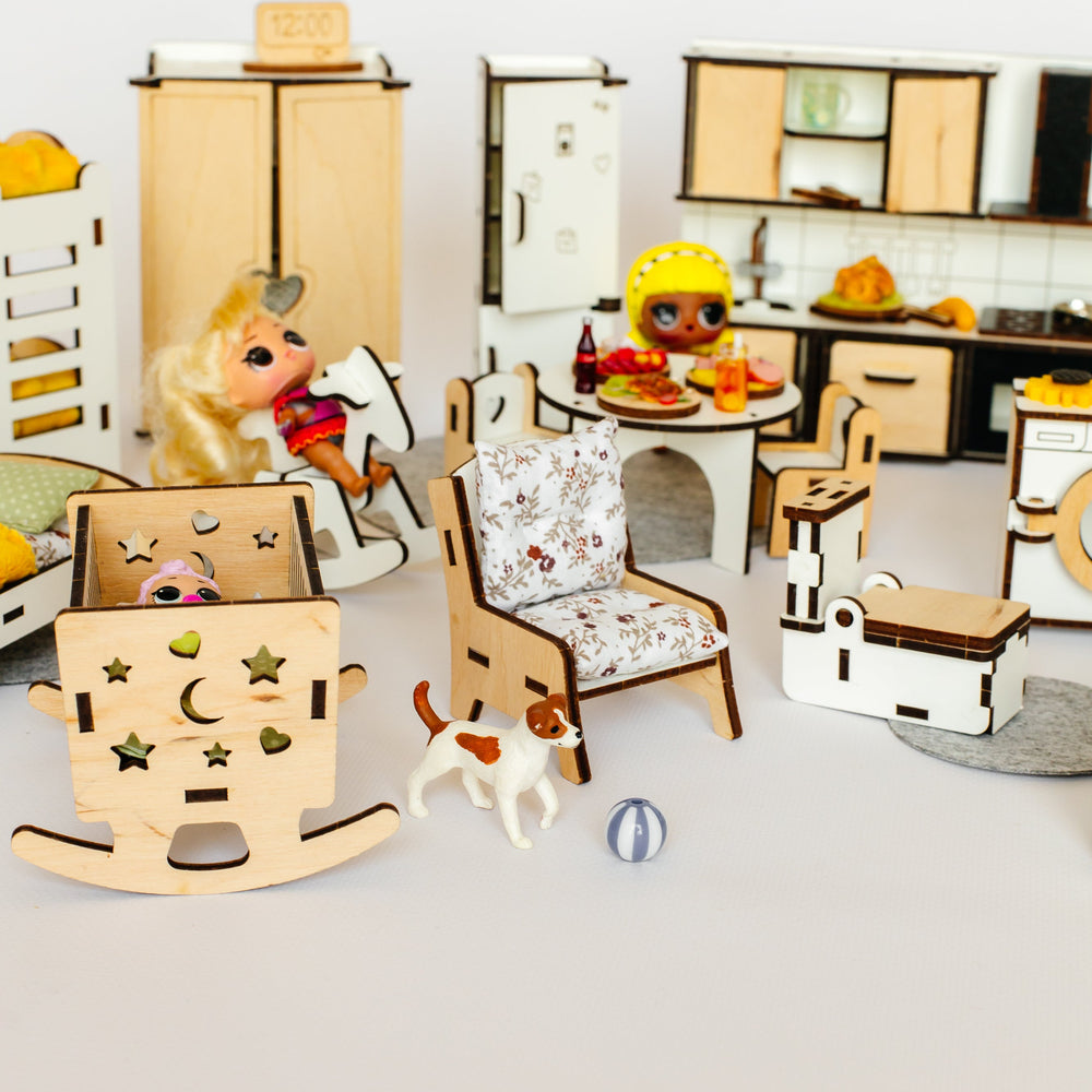 A miniature dollhouse-style interior scene with tiny furniture pieces, a small kitchen, a rocking chair with a doll seated in it, a patterned armchair, a toy cat, and other dollhouse accessories arranged on a light neutral surface.