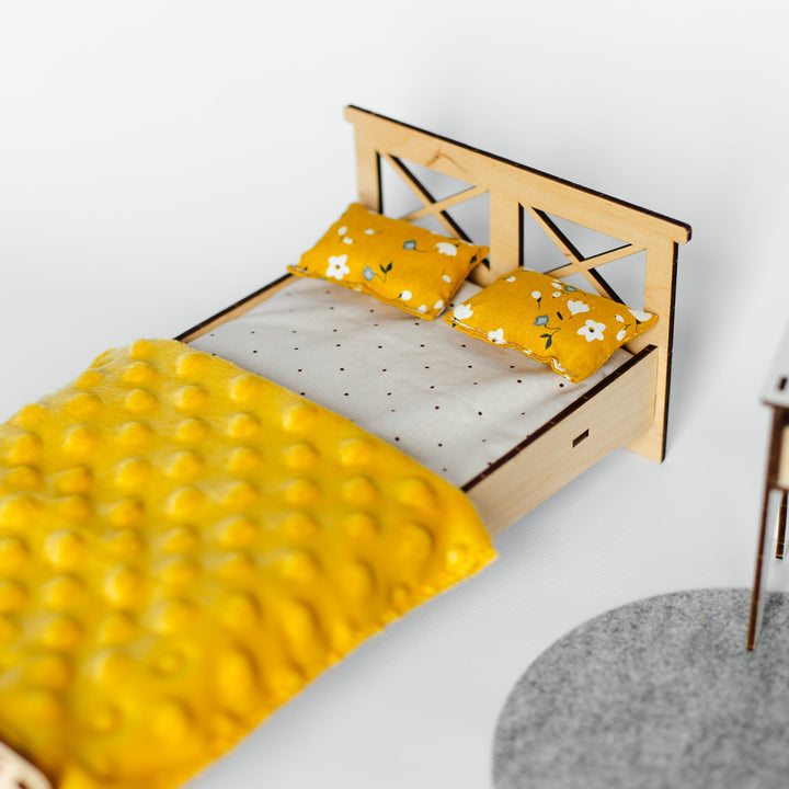 A miniature wooden bed frame with a yellow textured quilt and matching yellow pillows with white floral patterns, shown on a white surface next to a gray round rug.