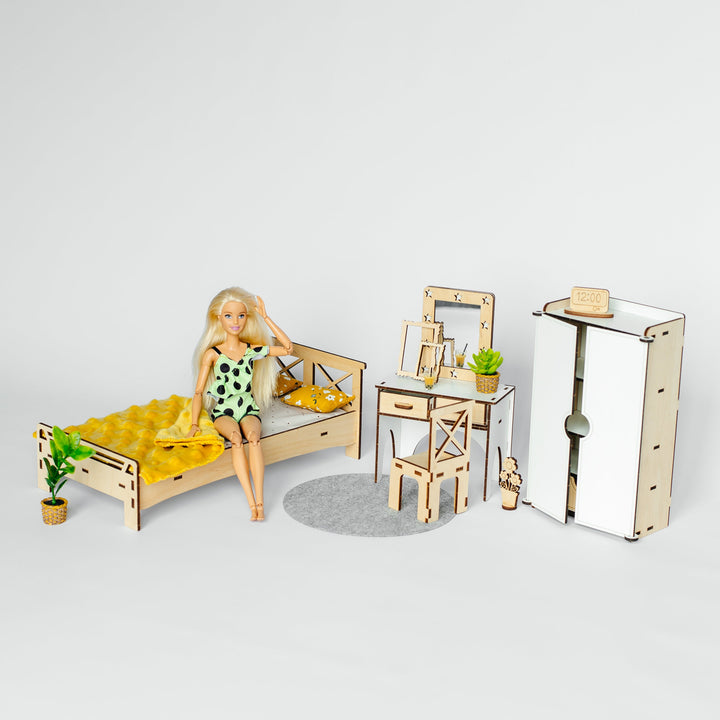 A dollhouse miniature bedroom set featuring a bed with yellow bedding, a doll sitting on the bed, a desk with a mirror and chair, and a tall wardrobe, displayed on a light gray background.