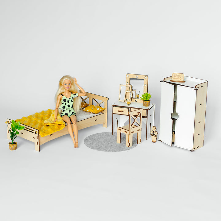 A miniature dollhouse bedroom set featuring a bed with a blonde doll wearing a green and white polka-dot dress, a wooden desk with a mirror, a wardrobe, and a small plant, all on a light gray background.