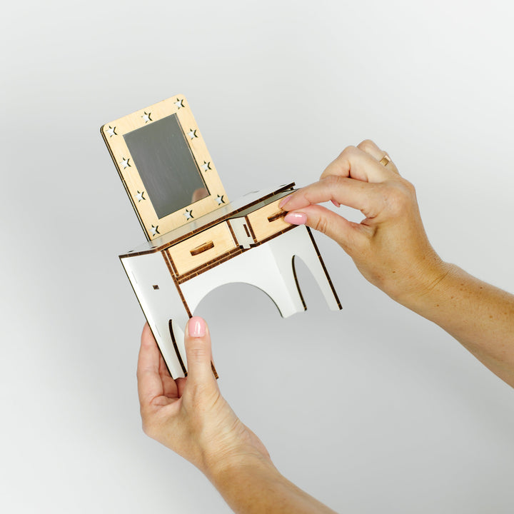 A person's hands holding a small wooden desk organizer with a mirror and drawers against a plain light gray background.