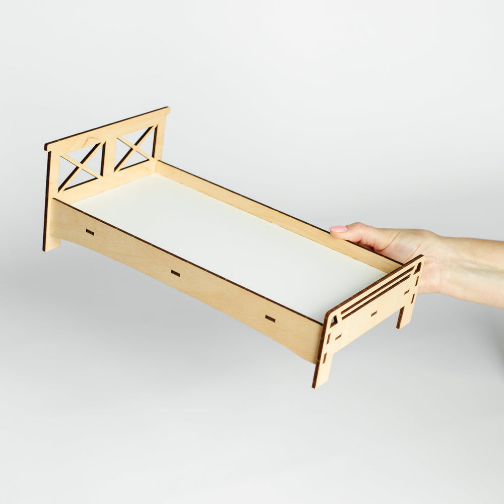 A miniature laser-cut wooden bed frame with a white mattress insert, held by a person's hand against a light gray background.