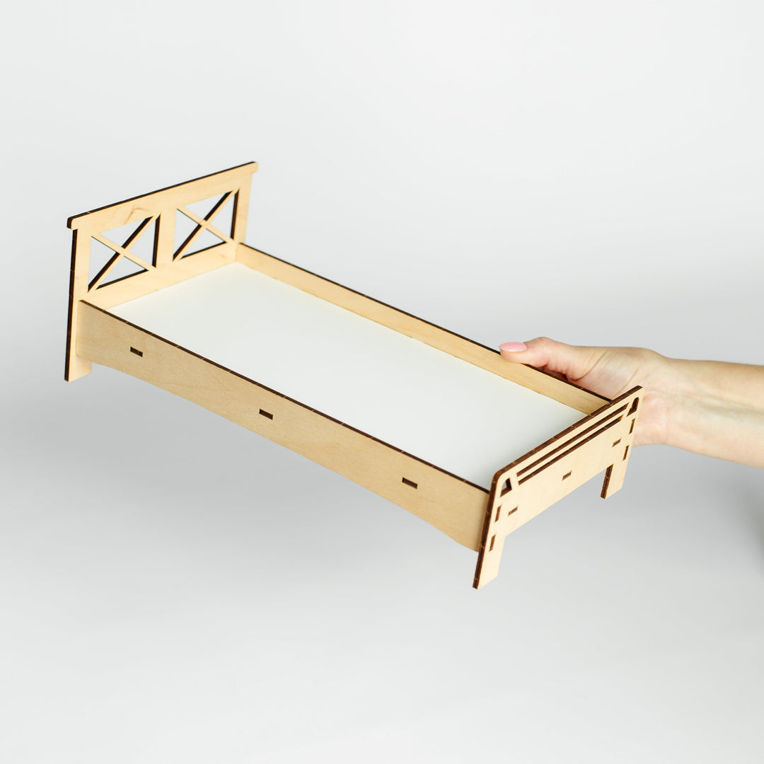 A miniature laser-cut wooden bed frame with a white mattress insert, held by a person's hand against a light gray background.