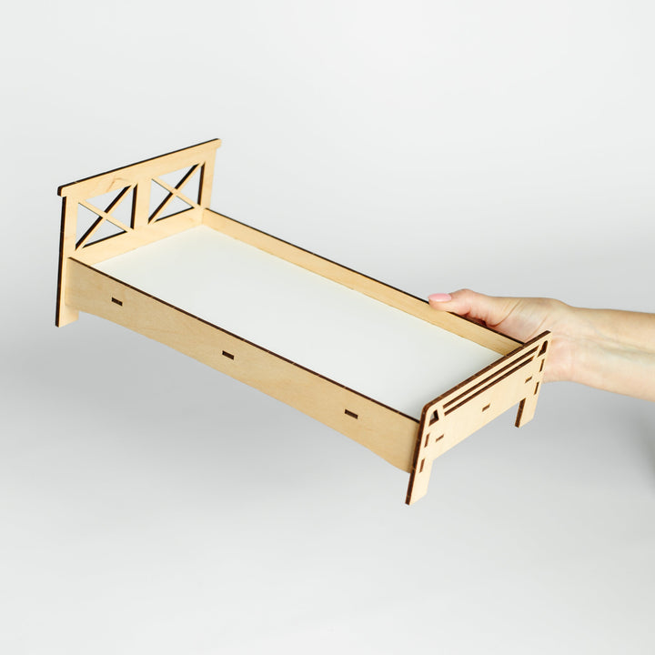 A miniature laser-cut wooden bed frame with a white mattress insert, held by a person's hand against a light gray background.
