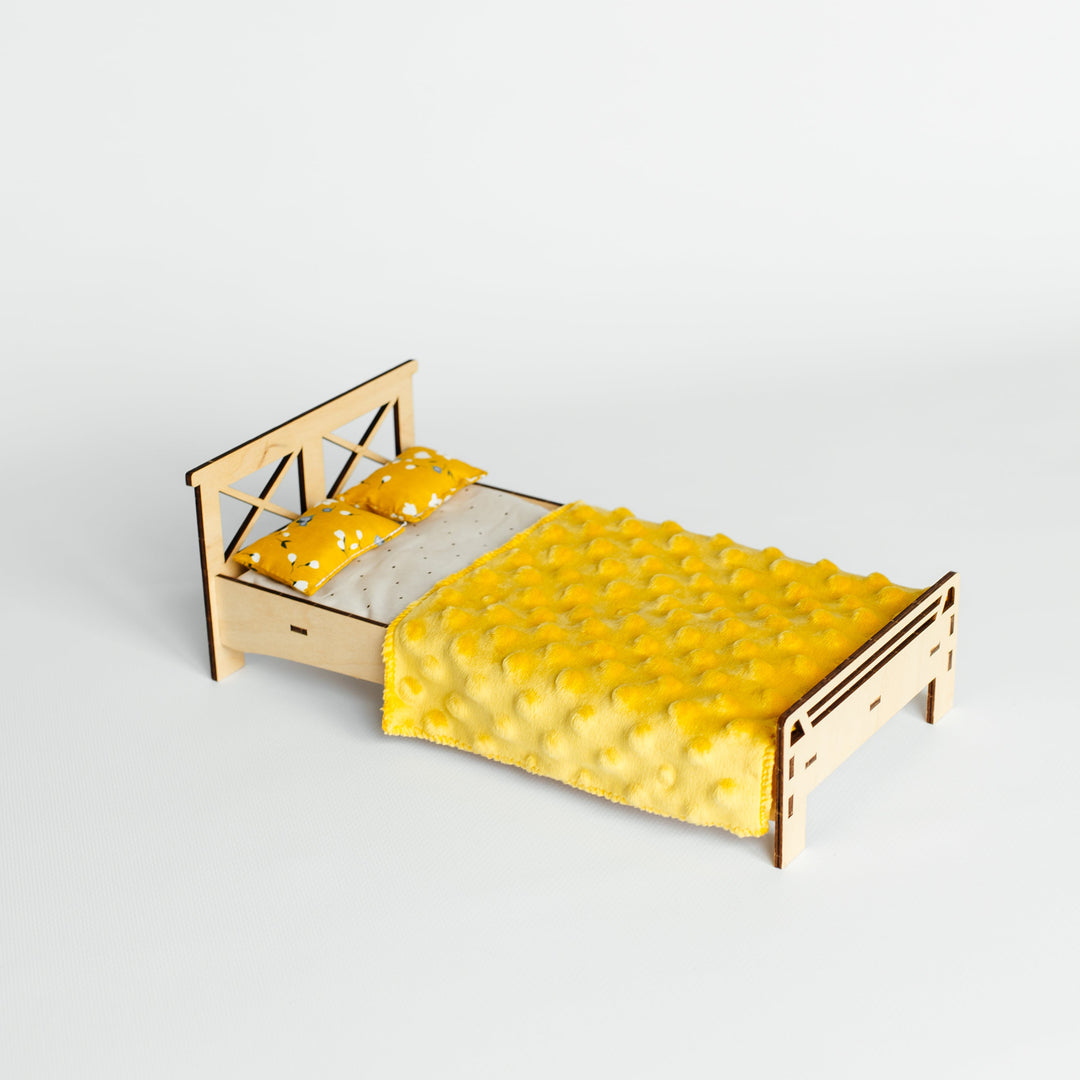 A miniature wooden bed frame with a geometric headboard and footboard, styled with a bright yellow tufted quilt and matching yellow pillows on a light gray background.