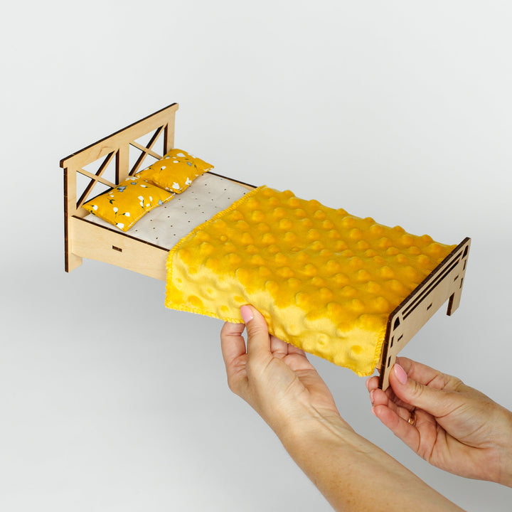 A miniature wooden bed frame with a yellow textured blanket and matching pillow, held by two hands against a plain light gray background.