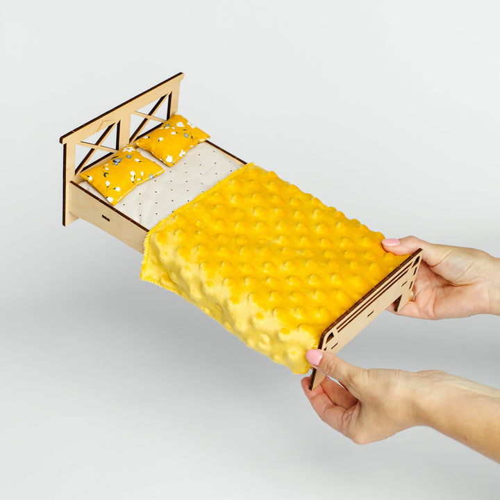A miniature wooden bed frame with a yellow textured blanket and matching pillows, held by two hands against a light gray background.