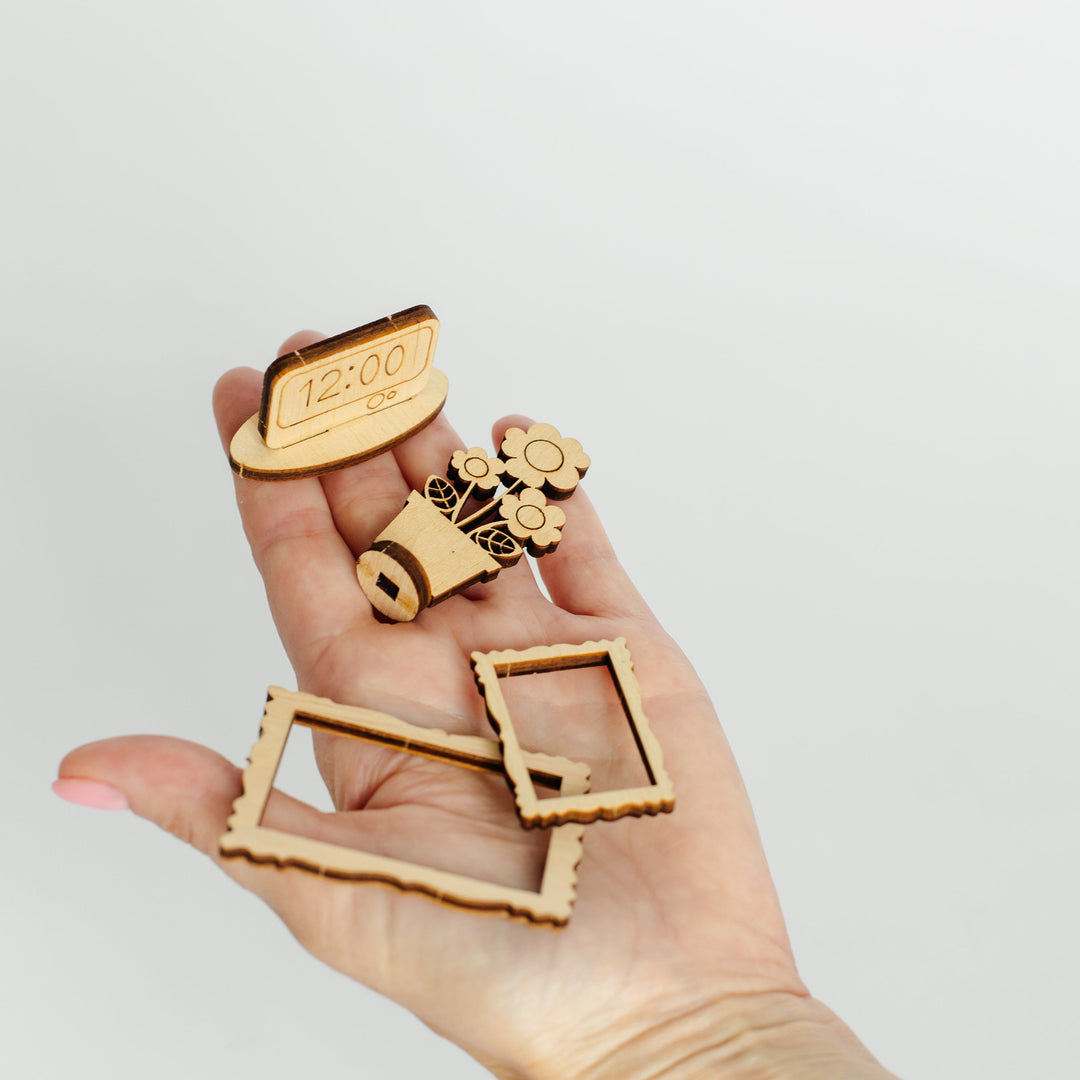A hand holding several small laser-cut wooden decorative pieces, including a clock face, gears, and rectangular frame-shaped cutouts, against a plain light gray background.