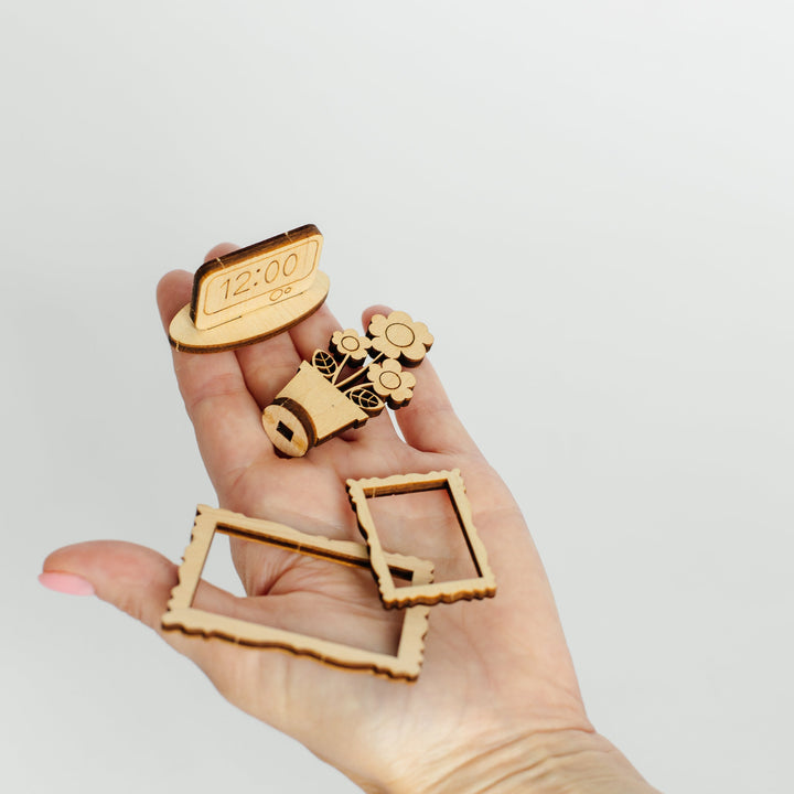 A hand holding several small laser-cut wooden decorative pieces, including a clock face, gears, and rectangular frame-shaped cutouts, against a plain light gray background.
