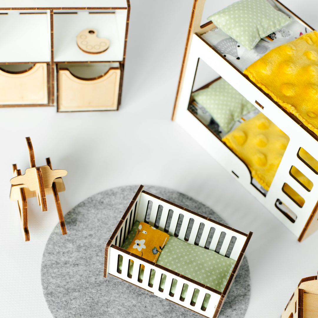 A miniature dollhouse crib with a green patterned blanket and yellow pillow, set on a gray felt rug. The scene is styled as a dollhouse with matching miniature furniture, including a changing table, storage boxes, and a rocking horse, on a white background.