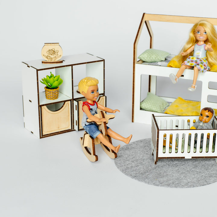 A dollhouse-style nursery scene with miniature furniture and dolls: a wooden crib with bedding, a small shelf with a plant and decorative item, and a rocking chair with a blonde doll seated in it. The set is photographed on a clean light gray studio background.