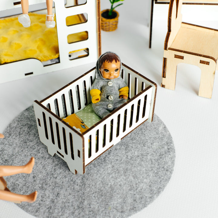 A miniature doll crib made of white and brown wood, containing a small doll, set on a round gray felt rug in a toy room scene.