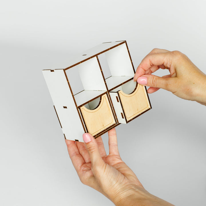 A pair of hands holding a small white and light wood modular organizer box against a plain light gray background.