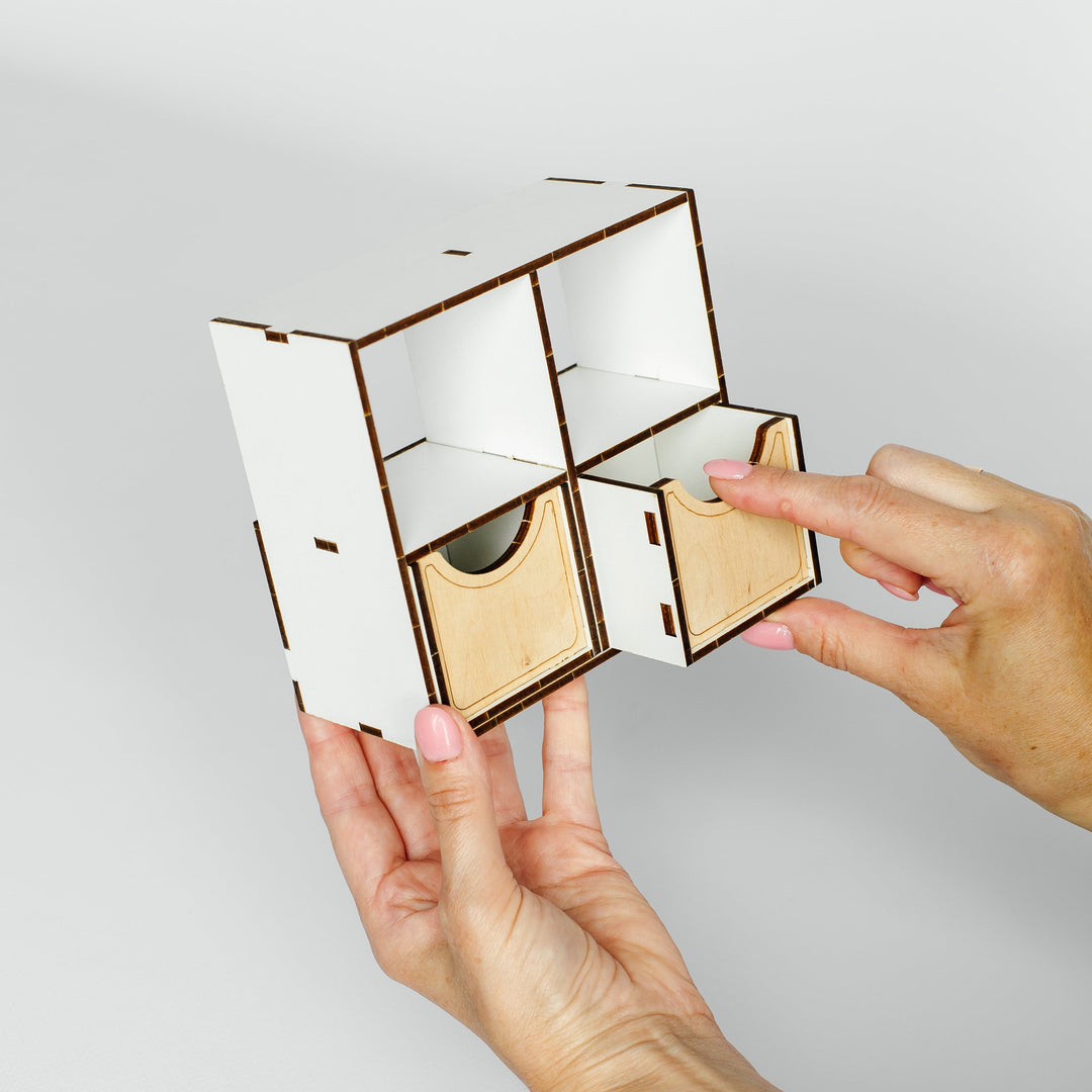 A person's hands holding and demonstrating a small white and light wood modular storage organizer with sliding compartments against a plain light gray background.