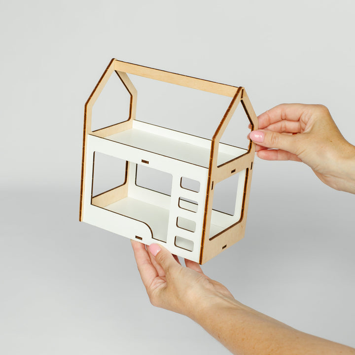 A person's hands holding a small, house-shaped wooden shelf or organizer with a white interior and light natural wood edges against a plain light gray background.