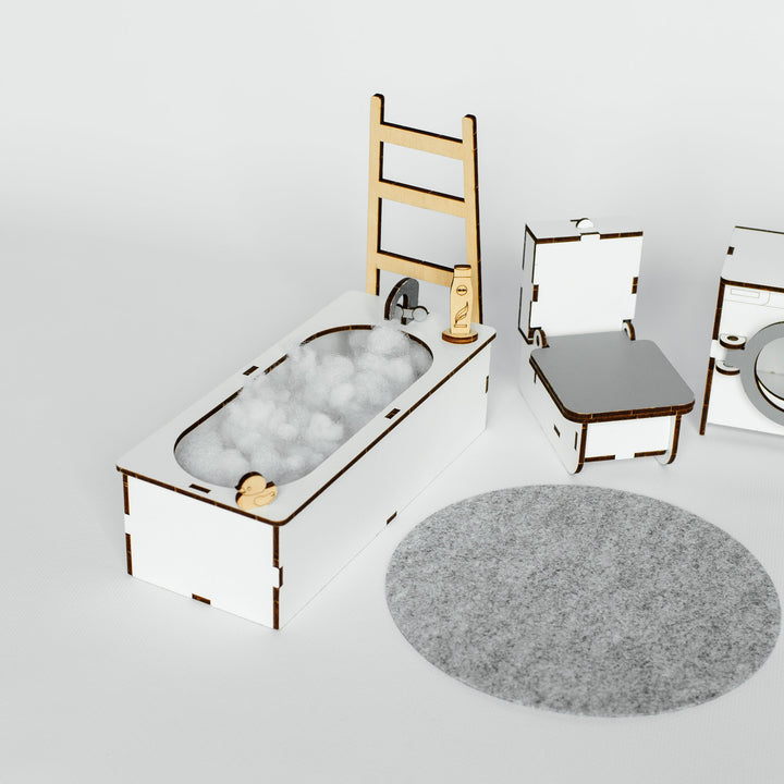 A miniature toy bathroom set featuring a white bathtub filled with white bath balls, a wooden ladder, a white toilet with a gray seat, a gray round rug, and a small white sink unit, all arranged on a light gray background.