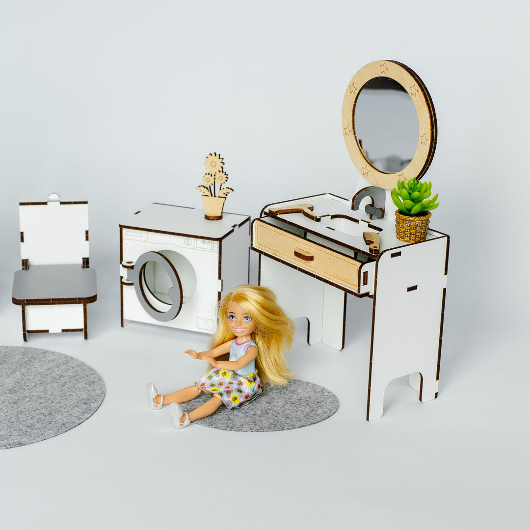 A miniature dollhouse-style room set with a blonde doll seated on a gray rug, including a white vanity with mirror, sink, and small plant, a washing machine, a chair, and two round rugs on a light gray background.