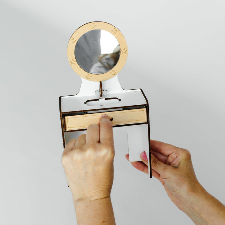 A person's hands holding a small wooden vanity table with a round mirror and a sliding drawer, set against a plain light gray background.
