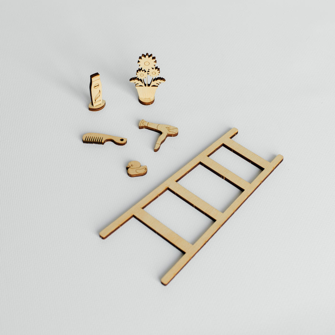 A miniature wooden ladder set with several small laser-cut accessories, including a comb, a small tool, a heart-shaped piece, and a flower-shaped piece, arranged on a light gray background.