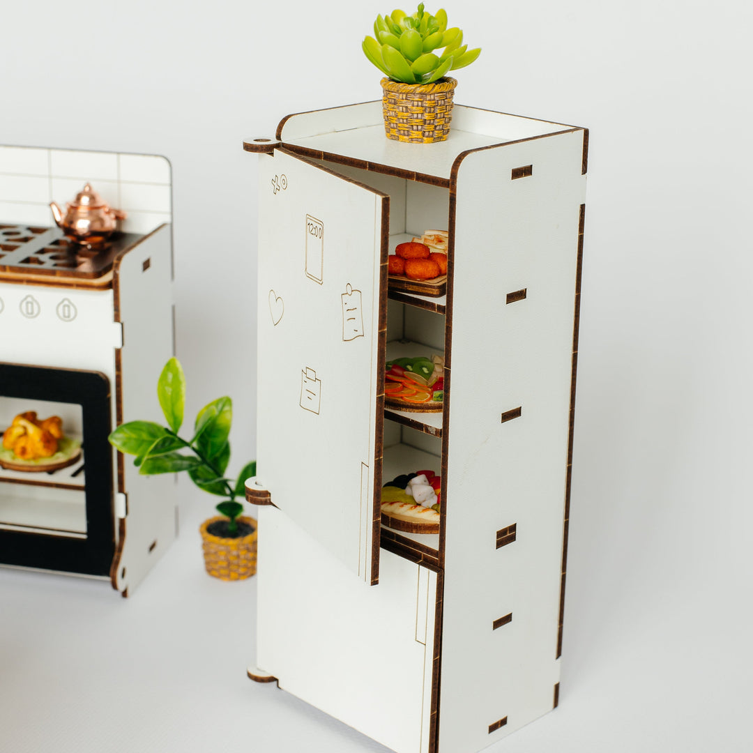 A miniature white laser-cut toy refrigerator with an open door showing small food items inside, alongside a small potted plant on top and another plant nearby, set against a plain light gray background.