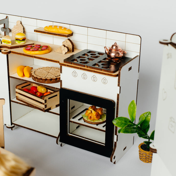 A miniature wooden toy kitchen set with a white and brown cabinet, black stovetop, shelves with toy food items, and a small potted plant on a light gray background.