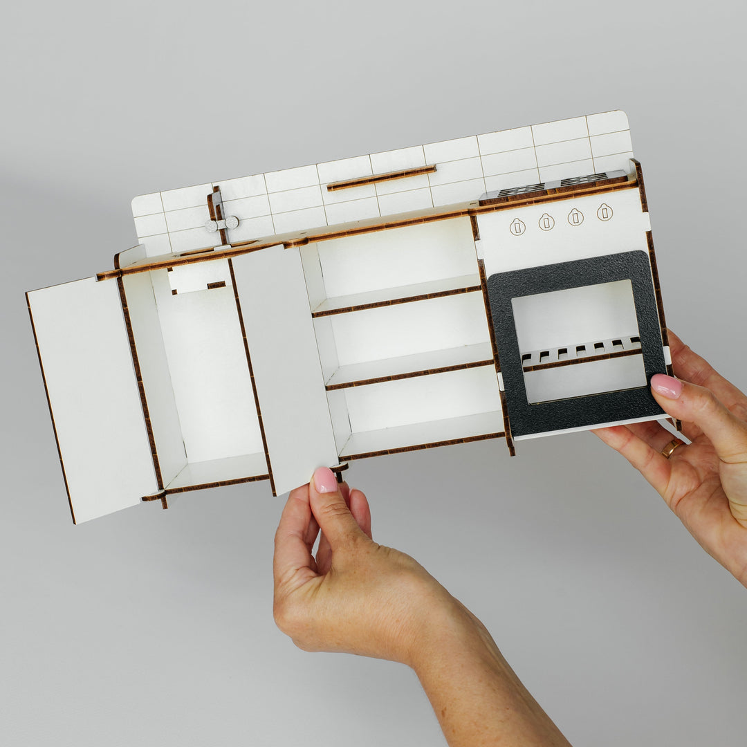 A miniature kitchen cabinet set with white tiles, brown wooden frames, and a black oven door, held open by two hands against a plain light gray background.