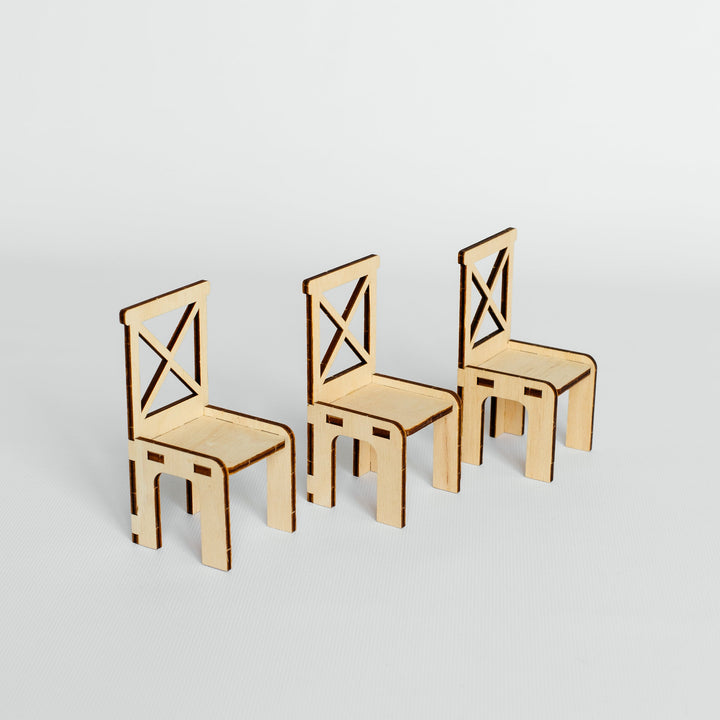 Three miniature laser-cut wooden chairs with a cross-back design, arranged in a row on a light gray background.