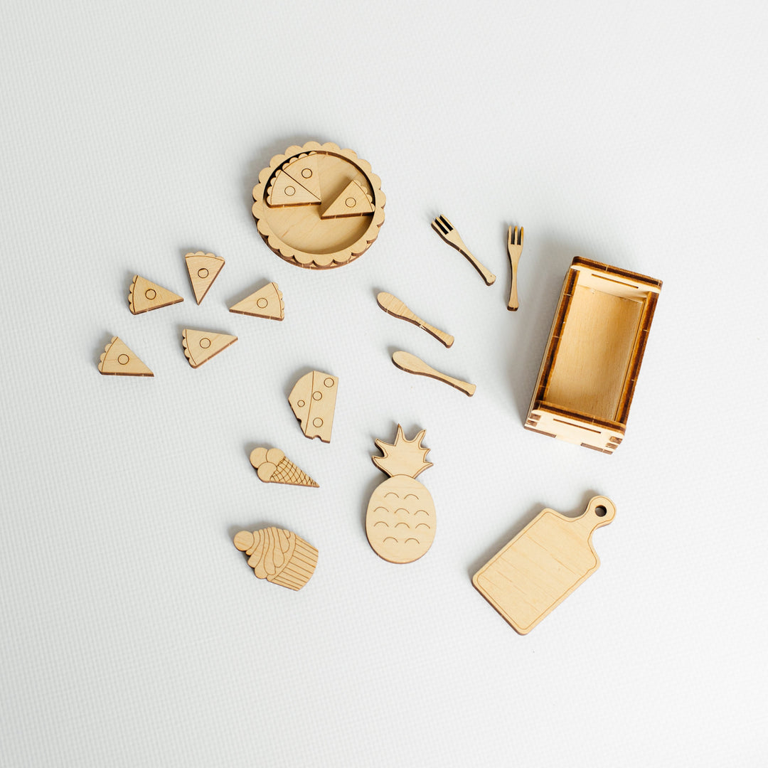 A wooden toy kitchen play set arranged on a light gray background, including a rectangular box, small utensils, and food pieces like pizza slices, an ice cream cone, a pineapple, and a cutting board.