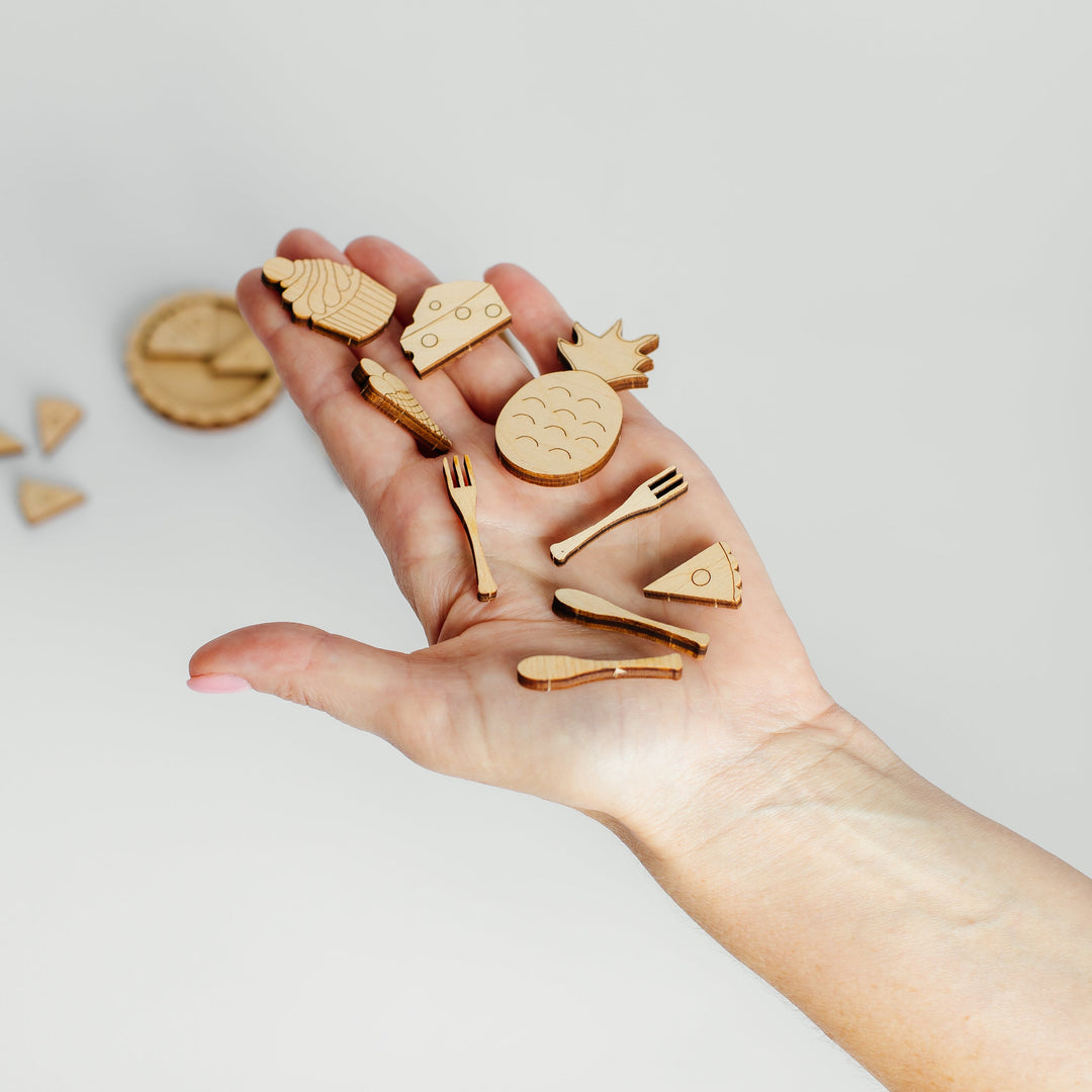 A hand holding several small wooden food-themed charms, including a pineapple, pizza slice, hamburger, fork, knife, and round piece, against a plain light gray background.
