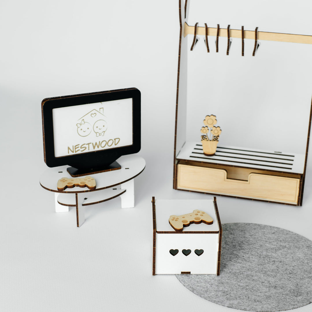 A miniature wooden toy furniture set featuring a TV stand with a TV displaying the 'NESTWOOD' logo, a small cabinet with a drawer, a clothing rack with hanging pegs, and a small box with heart cutouts, arranged on a light gray surface with a gray circular mat.
