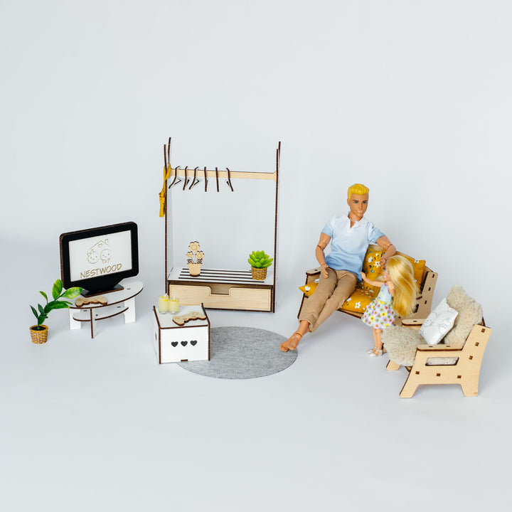 A miniature dollhouse living room set featuring a man and woman figurine sitting on a sofa, with a TV, side table, clothing rack, and other small furniture pieces arranged on a light gray background.
