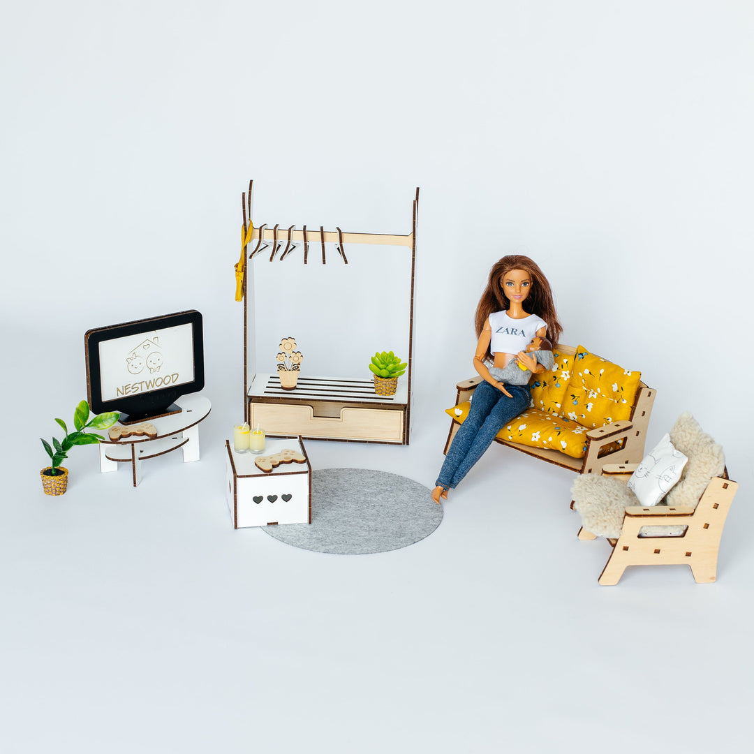 A dollhouse-scale furnished room set with a seated doll figure on a yellow patterned sofa, including a TV, cabinet, clothing rack, chair, rug, and plants, photographed on a clean white studio background.
