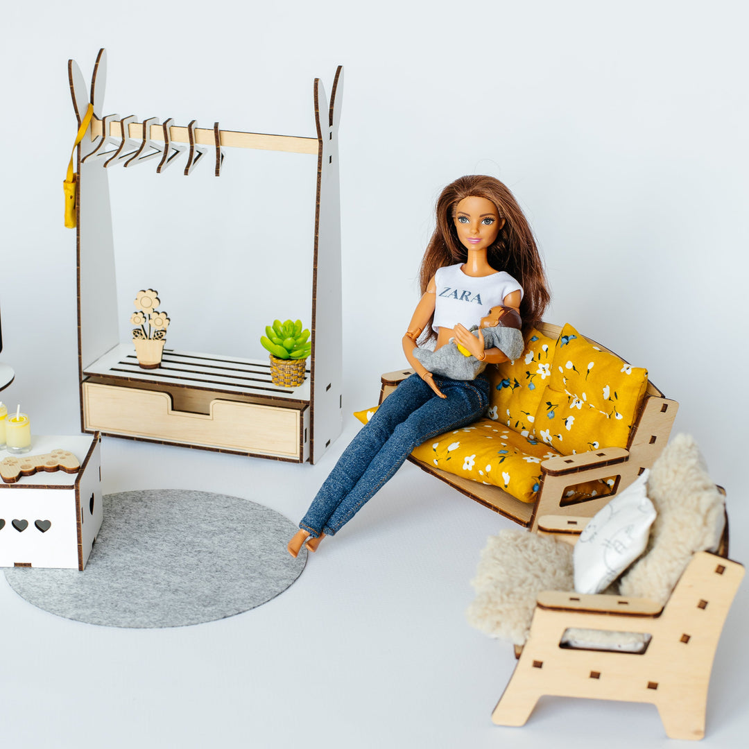 A doll sits on a small yellow floral sofa in a miniature room setup with a wooden coat rack, chair, rug, and storage box against a plain light background.