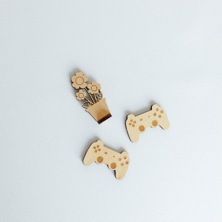 Three small wooden game controller-shaped accessories with floral details, arranged on a light gray textured background.