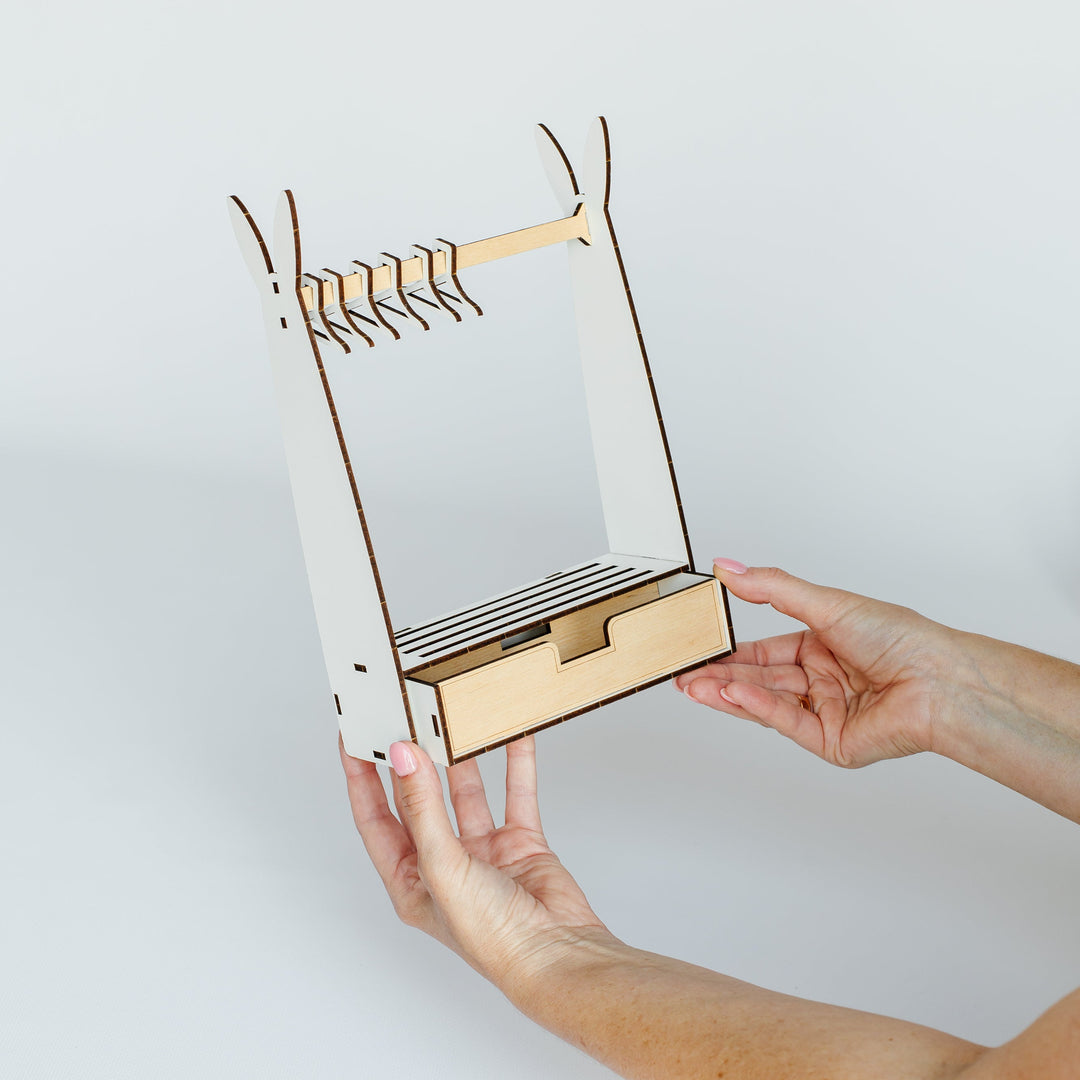 A person's hands holding a small wooden organizer with a drawer and hanging rod against a plain light gray background.