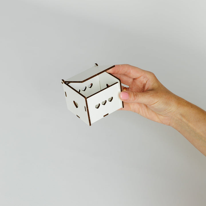 A hand holding a small white laser-cut cardboard box with brown edges against a plain light gray background.