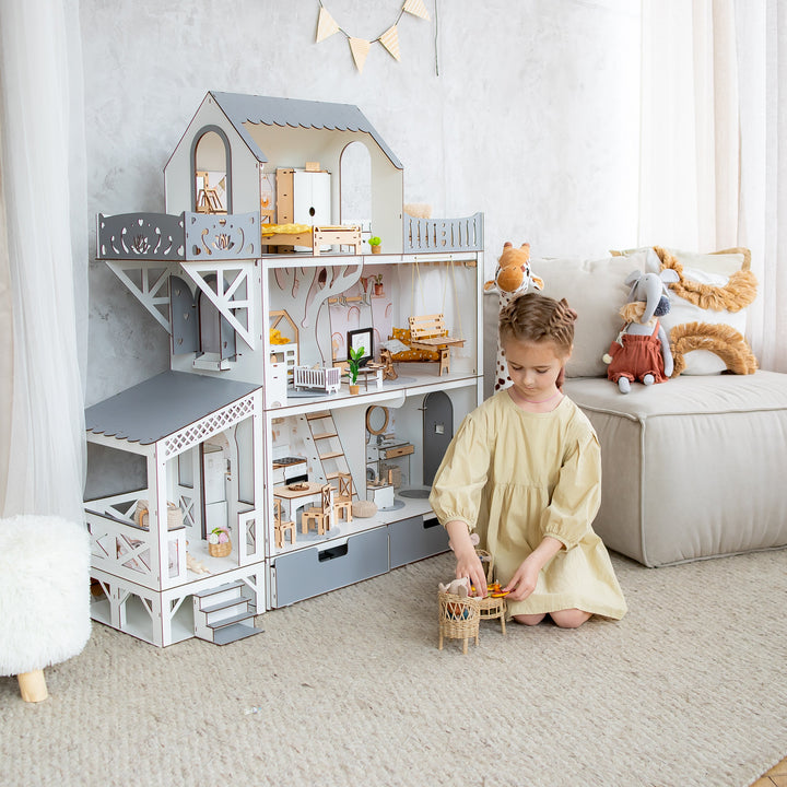 A young child in a cream dress sits on a rug in a bright room, playing with a miniature dollhouse. The dollhouse is a large, multi-level white and gray house filled with tiny furniture pieces.
