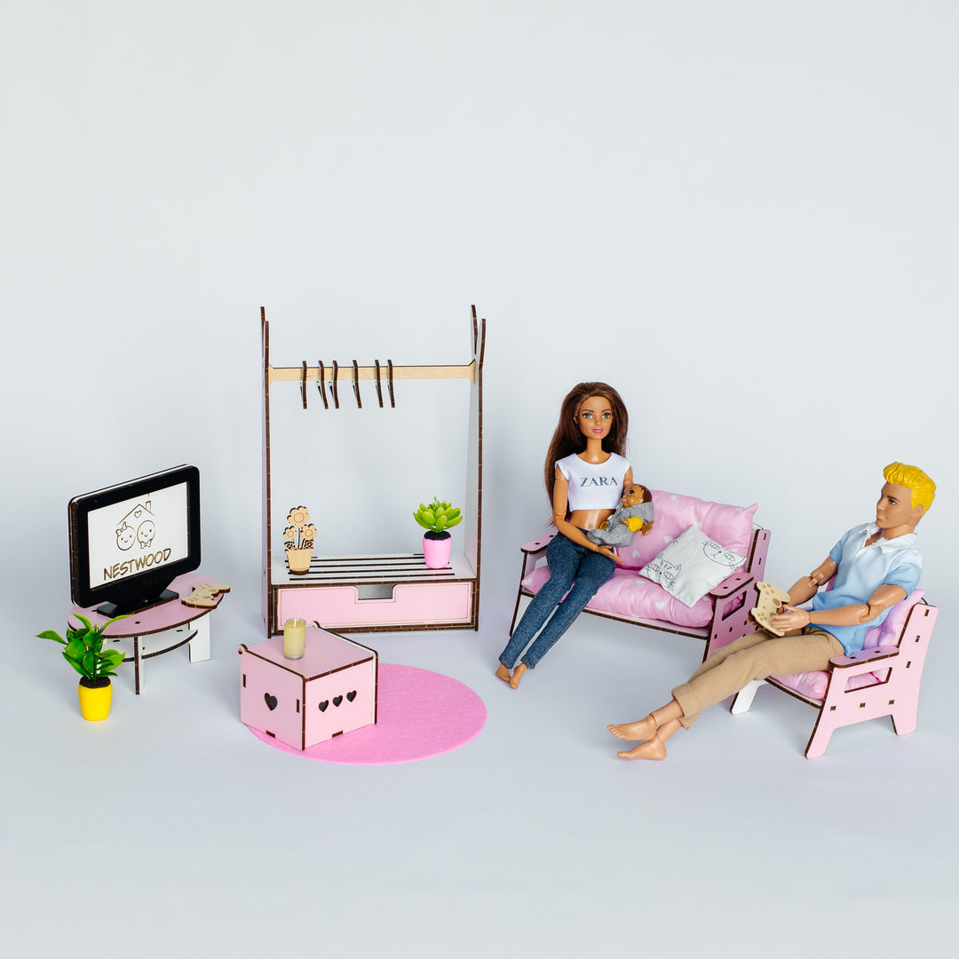 A miniature dollhouse living room set featuring a pink sofa with a female doll sitting on it, a pink chair with a male doll reclining, a pink coffee table, a clothing rack, and a small TV on a stand, all arranged on a light gray background.
