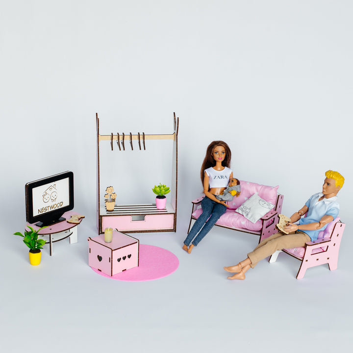 A miniature dollhouse living room set featuring a pink sofa with a female doll sitting on it, a pink chair with a male doll reclining, a pink coffee table, a clothing rack, and a small TV on a stand, all arranged on a light gray background.