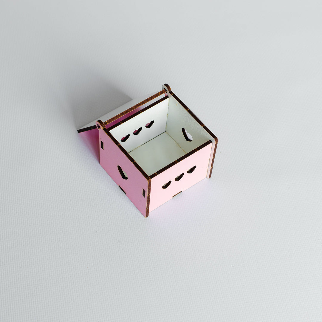 A small pink wooden box with a hinged lid, featuring laser-cut eye and mouth details on the front, set against a plain light gray background.