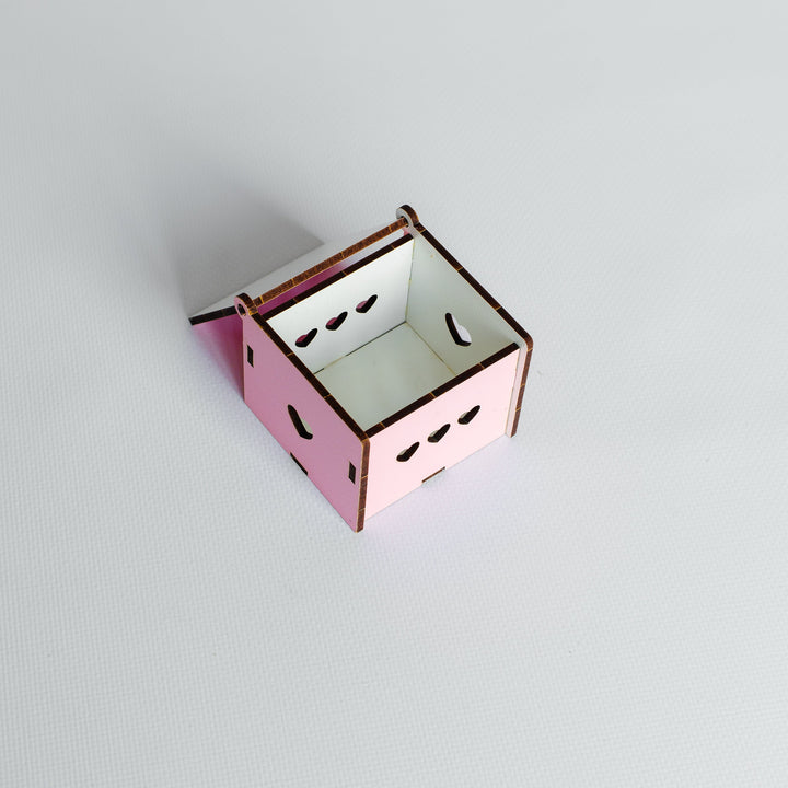 A small pink wooden box with a hinged lid, featuring laser-cut eye and mouth details on the front, set against a plain light gray background.