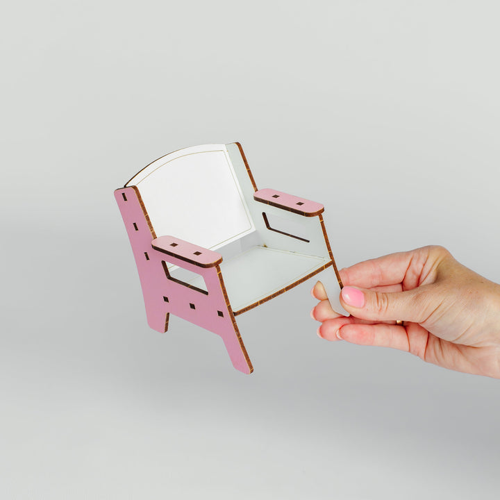 A small wooden chair with a pink frame and white seat, held by a person's hand against a light gray background.
