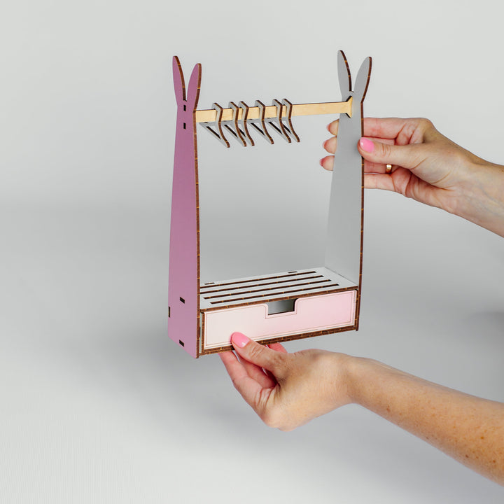 A person's hands holding a small wooden organizer shaped like a rabbit, featuring a pink body, gray ears, a wooden hanger bar with metal clips, and a small drawer compartment.