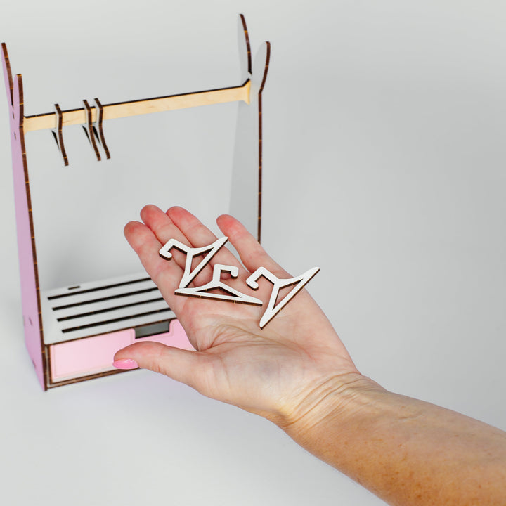 A hand holding a small white geometric accessory in front of a pink and brown miniature clothing rack model, set against a plain light gray background.