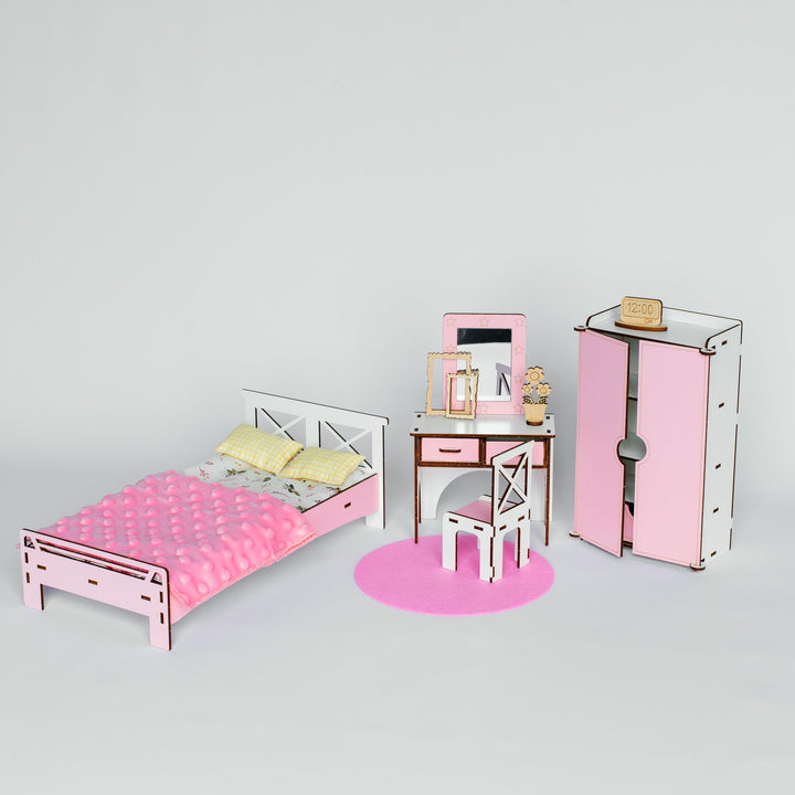 A dollhouse-style bedroom furniture set in pink and white, including a single bed with pink bedding, a desk with chair, a tall wardrobe, and a round pink rug, photographed on a light gray studio background.