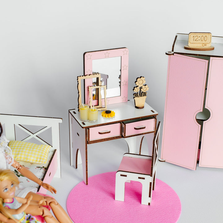 A dollhouse miniature vanity set with a pink and white desk, matching chair, mirror, and accessories, displayed alongside a pink wardrobe and bed with dolls in a dollhouse room.